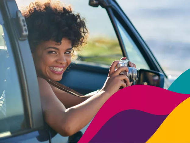 A happy woman holding a camera looks out of her vehicle as she goes on a travel adventure.